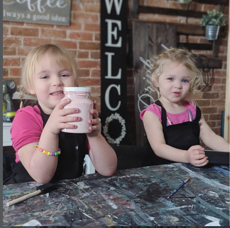 Two small children in aprons at a paint-covered workshop table